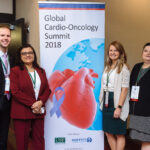 A man and three women pose in front of a banner for the Global Cardio-Oncology Summit in 2018