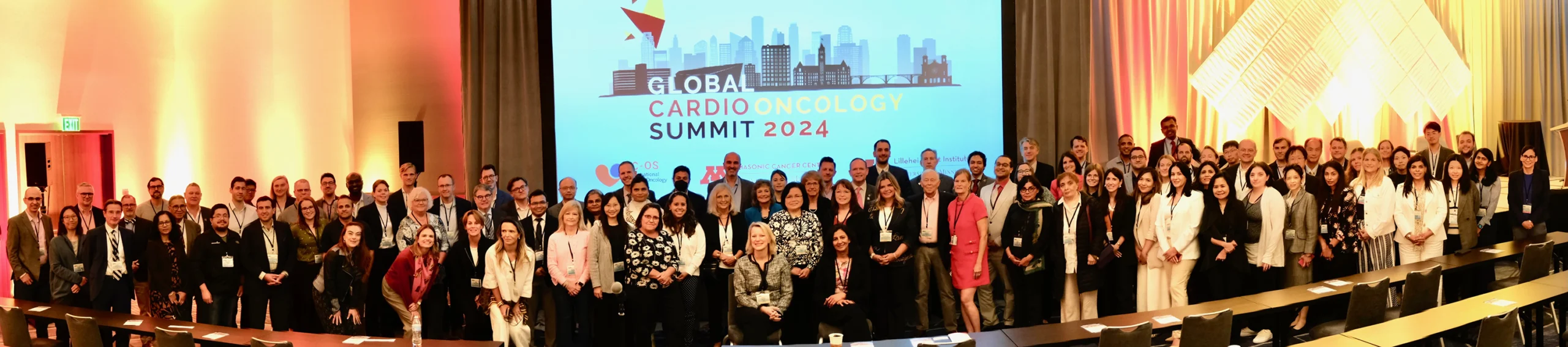 The attendees of the Global Cardio-Oncology Summit (GCOS) stand in front of a projector screen with the event logo