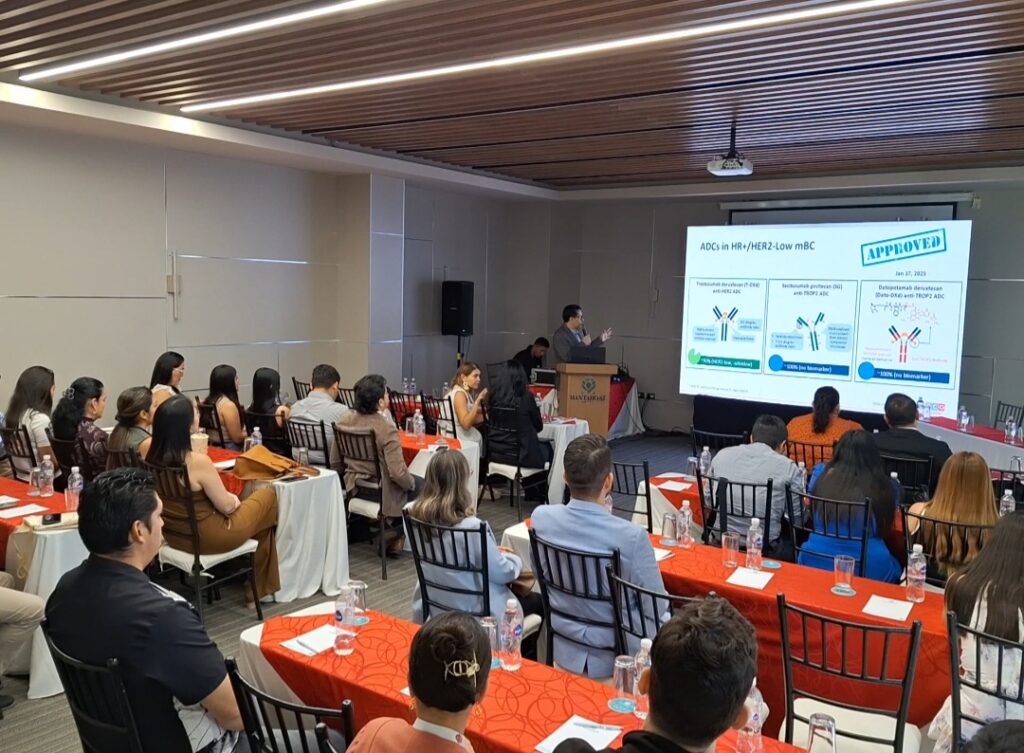 A group of cardio-oncologists sit a tables with orange table clothes while a person in the back of the room presents charts from behind a wooden podium