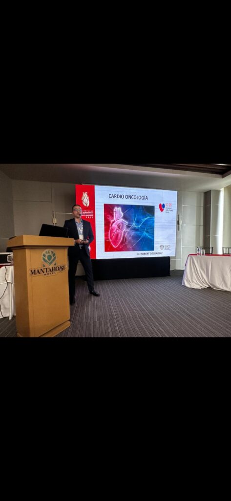 A mean in a dark suit stands to the right of wooden podium presenting cardio-oncology research on a projector screen