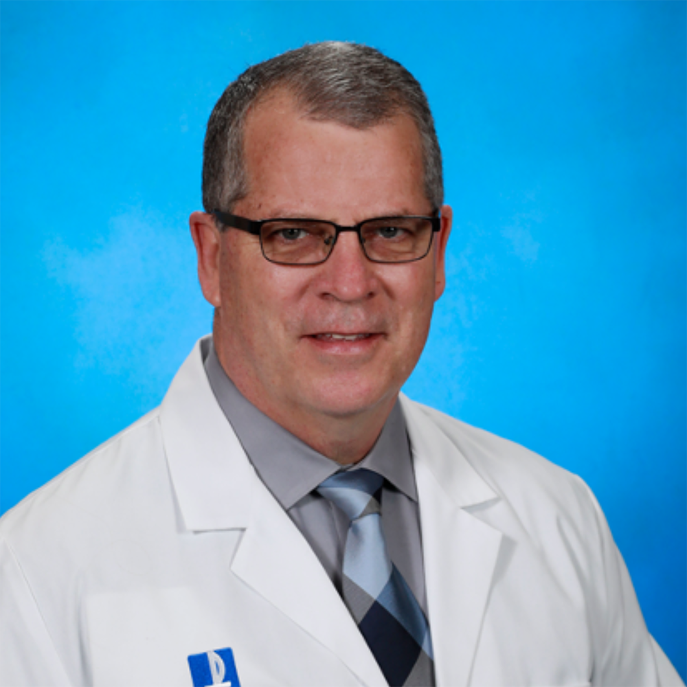 Dan Lenihan a white man with short grey hair and glass sits in a white lab coat in front of a blue background